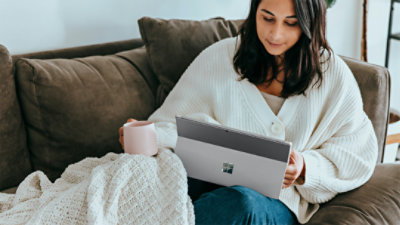 A woman sitting on a couch with a coffee cup and blanket, looking at a laptop