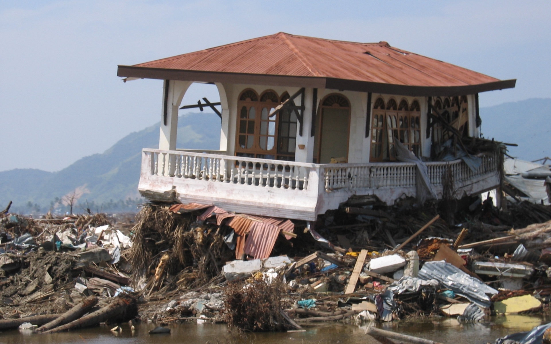 A white house with a red roof sits perched on a pile of debris above mud and water