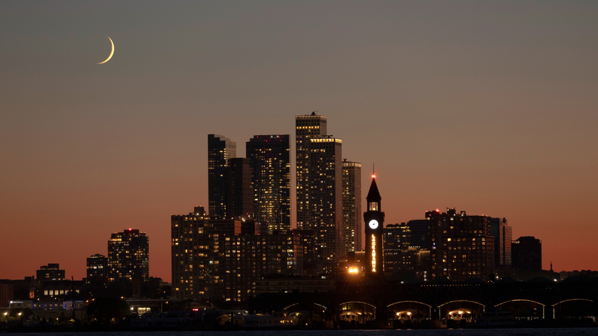 A yellow crescent moon is pictured above a city skyline. Skyscrapers are clustered in the middle of the screen with a body of water at the bottom.
