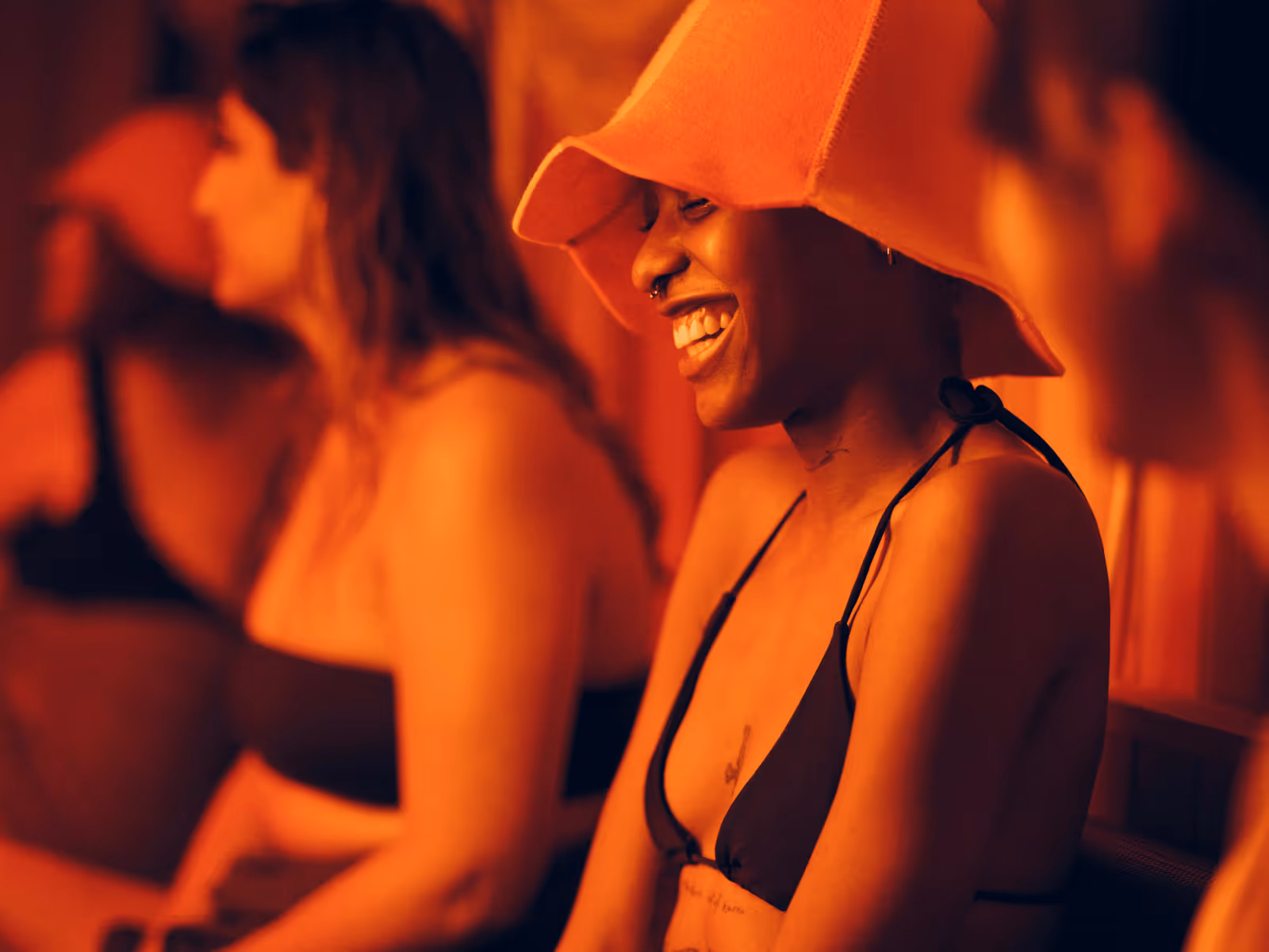 A smiling person wearing a large hat and black bikini top sits in a warmly lit, orange-toned room, surrounded by others enjoying a relaxed, communal atmosphere.