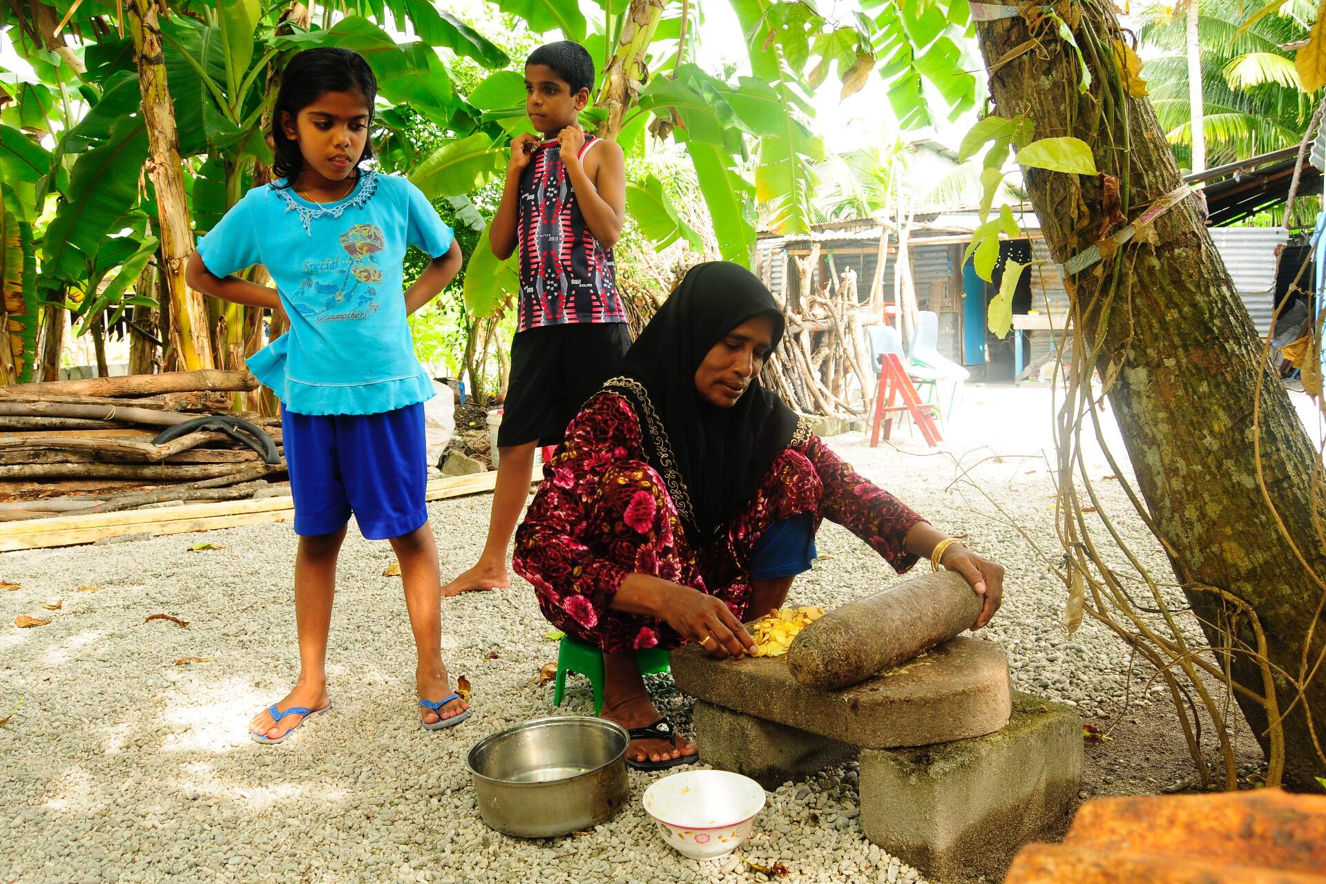 Two children are watching as a woman prepare food under the shadow of a tree.