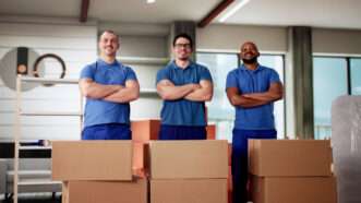 Three moving men in blue uniform shirts stand in a residential living room with arms crossed, in front of cardboard boxes. | Andrey Popov | Dreamstime.com