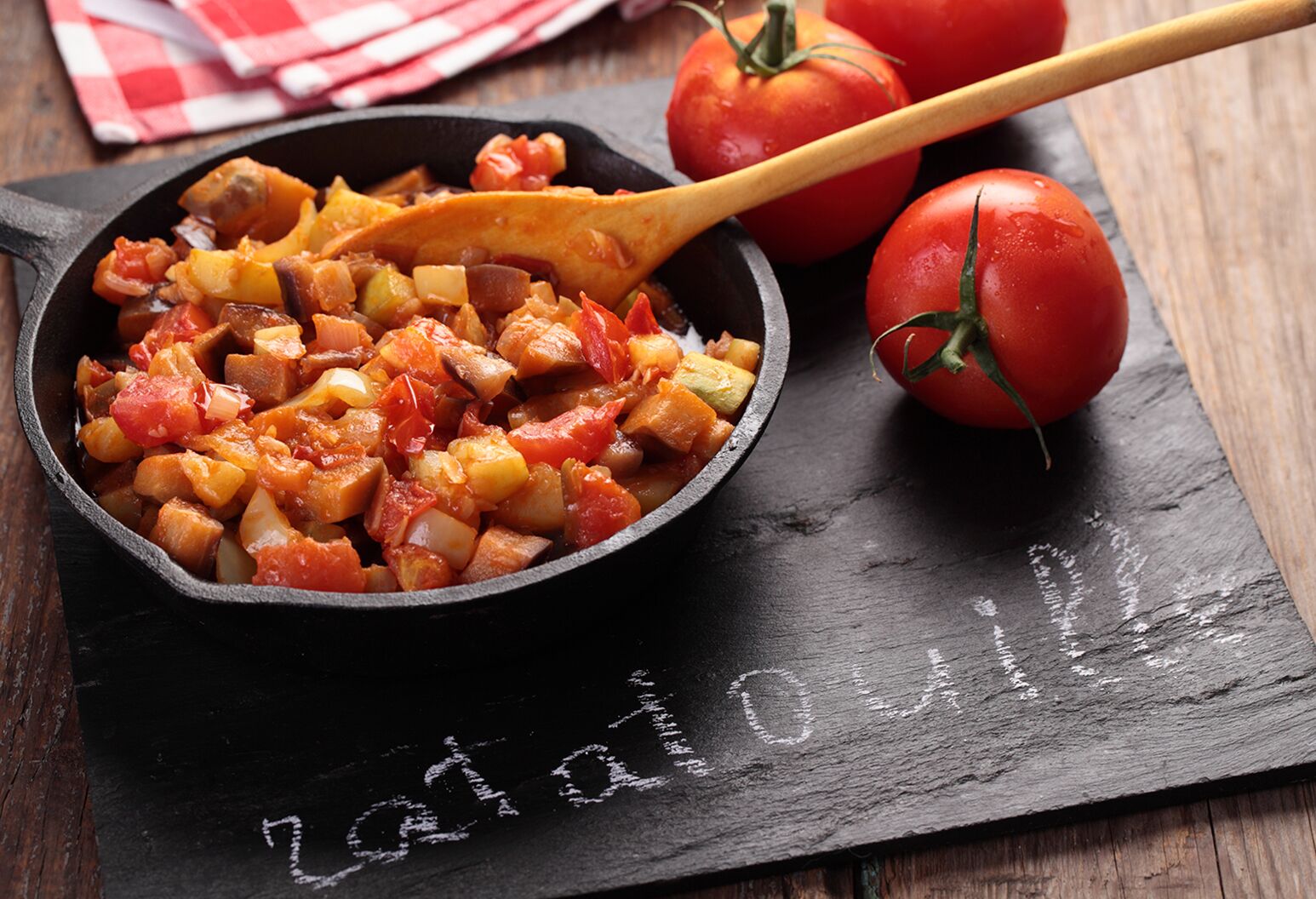 A bowl of ratatouille with a wooden spoon on a black cutting board with tomatoes nearby.