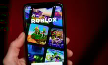 A hand holds a phone showing the Roblox homepage.