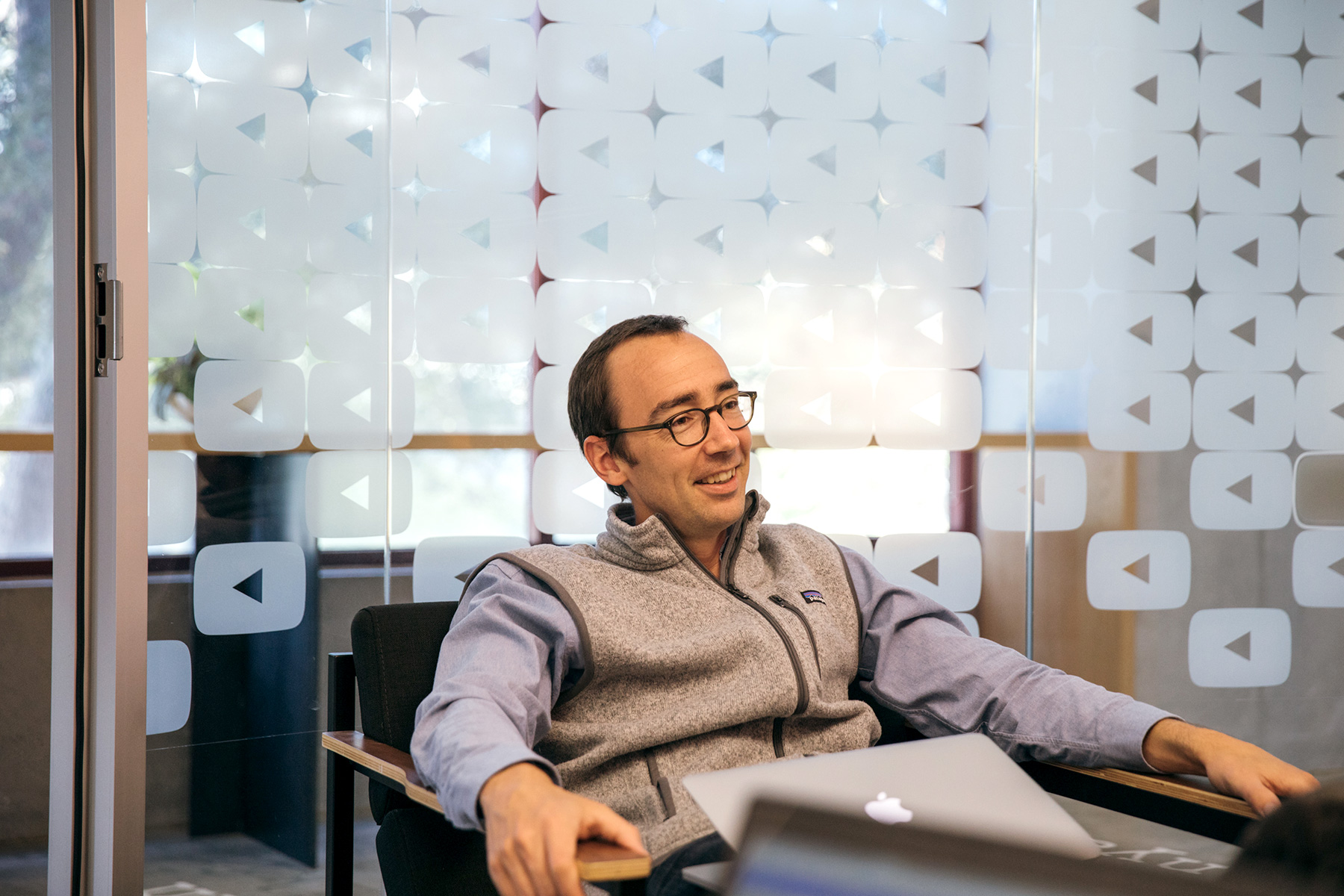 A man in a conference room with the YouTube logo on the wall, smiling and sitting in a chair.